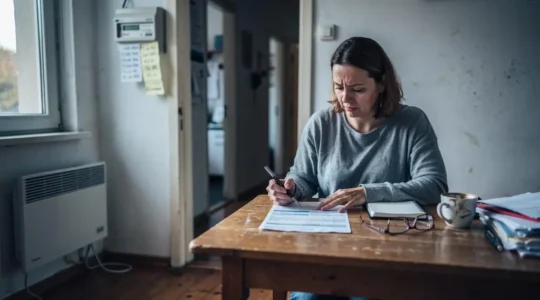 Une femme compare sa facture d'électricité avec ses relevés de compteur notés sur un carnet, dans le salon de son petit appartement.